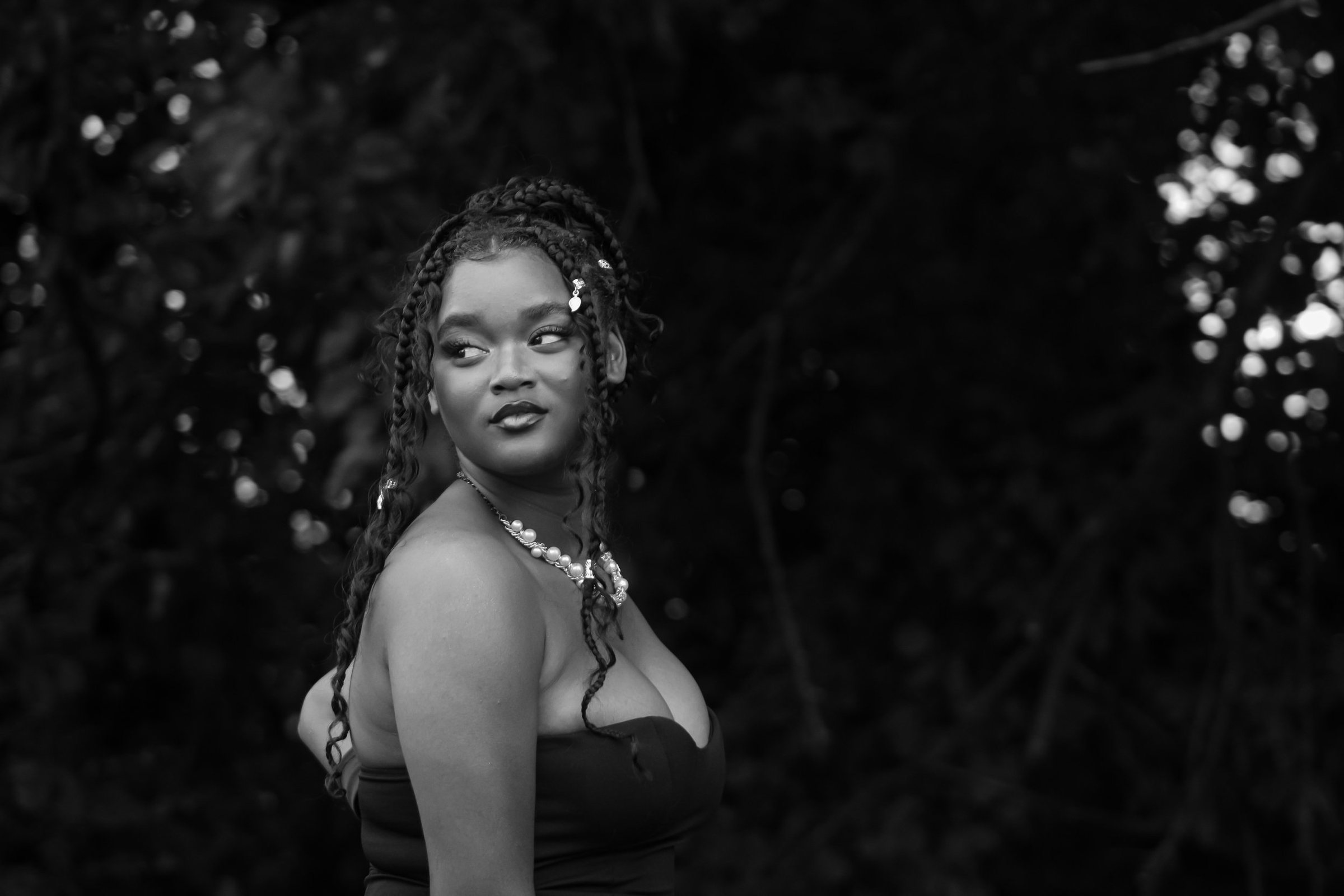 A black and white portrait by Leyna: braids with a pearl clip, a pearl necklace and a strapless dress, looking back over her shoulder among dark leaves